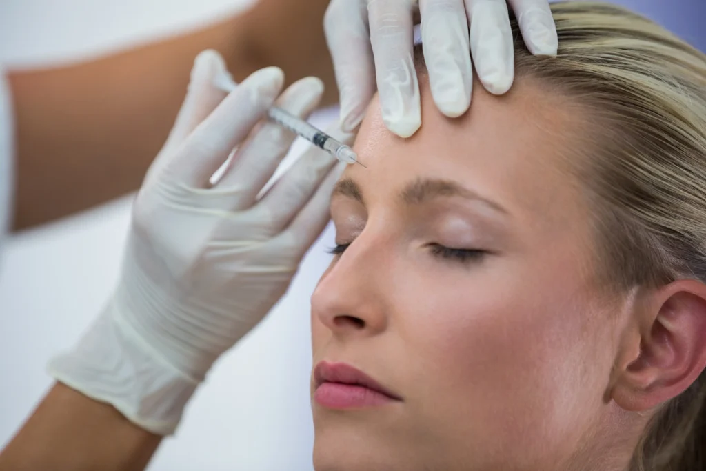 A close-up of a female patient with closed eyes receiving a Dysport injection on her forehead, administered by a medical professional wearing gloves.