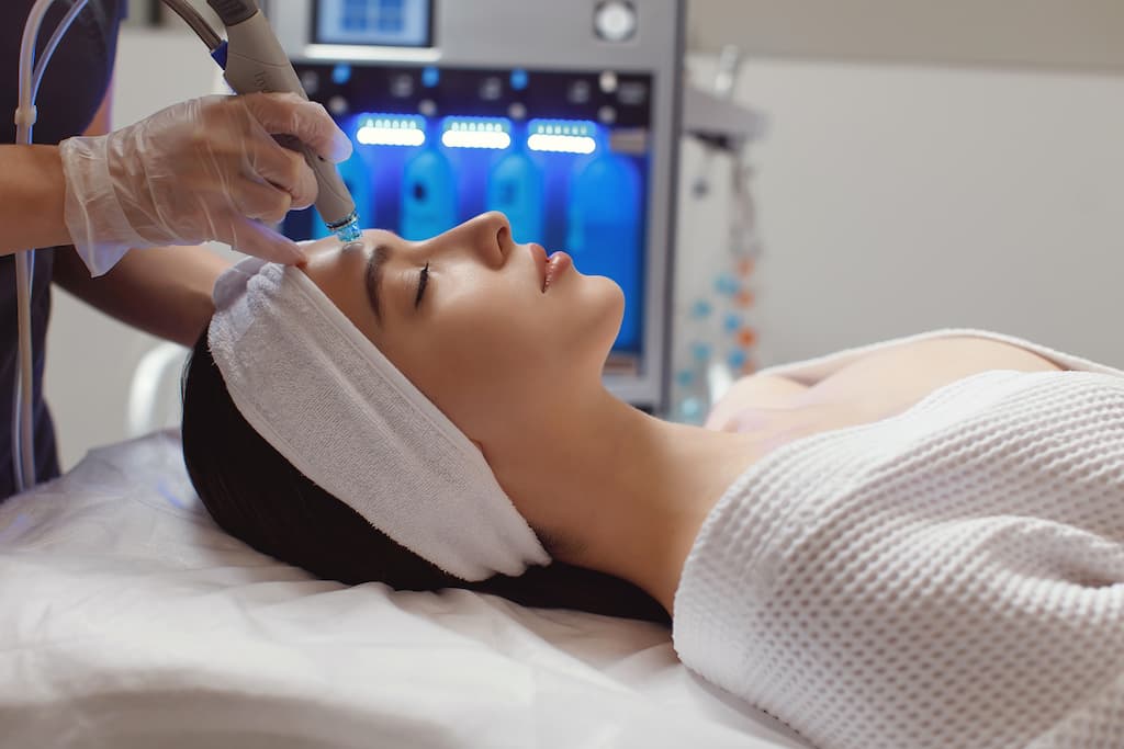 Woman receiving HydraFacial treatment lying on spa bed with practitioner using handheld device on her face, blue LED light therapy equipment visible in background